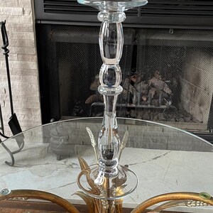 May include: A clear glass candlestick holder with a round top and a decorative base. The base has gold-colored metal leaves and wheat stalks. The candlestick holder is on a glass table.