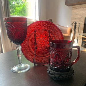 May include: A collection of ruby red glassware and a plate with a detailed pattern. The set includes a wine glass, a large plate, and a mug. The items are arranged on a dark surface, with natural light illuminating the scene.
