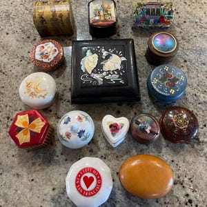 May include: A collection of 13 small decorative boxes with various designs and colors. The boxes are made of different materials, including wood, metal, and ceramic. Some of the boxes have lids, while others are open. The boxes are arranged in a random pattern on a light brown surface.