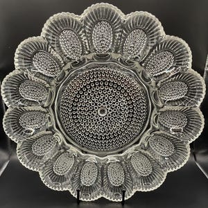 May include: Clear glass serving platter with a daisy-like design. The platter features a central circle and petal-shaped sections, each adorned with small, raised dots. The edges have a scalloped, textured pattern. The platter is displayed on a black stand.