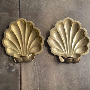 May include: A pair of matching brass wall sconces shaped like scalloped seashells. Each sconce features a polished gold-toned metal finish with deep, radiating ridges and a curved candle holder cup at the base, designed for mounting on a flat surface.