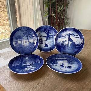 May include: A collection of blue and white porcelain plates with various landscape scenes. The plates are round and oval, featuring detailed illustrations of trees, animals, and buildings. The plates are displayed on a wooden table.