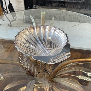 May include: A silver-colored, shell-shaped dish with a fluted design. The dish has a scalloped edge and sits on a glass table. The dish is likely used for decorative purposes or to hold small items.
