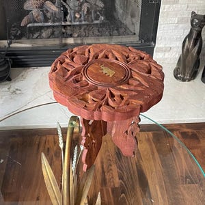 May include: A round, reddish-brown wooden plant stand featuring intricate floral carvings and a central circular brass inlay depicting an elephant. The stand is supported by three carved wooden legs and rests atop a metal base shaped like leaves.