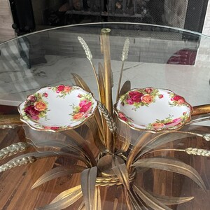 May include: Two white, scalloped-edge dishes with a floral rose pattern in red, pink, and orange. The dishes have a gold trim. They are displayed on a gold-colored decorative table with a glass top.