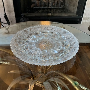May include: A clear, round glass serving platter with an intricate floral and starburst pattern. The edges are scalloped, and the platter sits on a gold-colored metal base. The glass is approximately 12 inches in diameter.