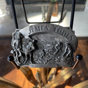 May include: A rectangular silver-toned metal belt buckle featuring a relief scene of a person with a donkey and figures mining in a river. The text AMES TOOLS is embossed at the top, and the text the Gold Rush of 1849 is embossed at the bottom.