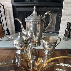 May include: A three-piece silver-plated tea set consisting of a tall coffee pot with a lid, a sugar bowl with a lid, and a creamer. Each piece features a polished metallic finish and a decorative embossed band with floral details around the rim.