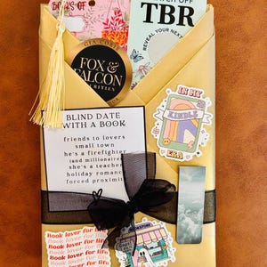 May include: A gold-wrapped book package featuring a black ribbon bow, a 'Blind Date with a Book' card, a 'Scratch Off TBR' card, a 'Books of' bookmark with a yellow tassel, a 'Fox & Falcon' sticker, and various decorative stickers including 'In My Kindle Era'.