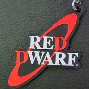 Könnte beinhalten: Ein schwarzer und roter Schlüsselanhänger mit dem Text "RED DWARF" in weißen Buchstaben. Der Schlüsselanhänger hat eine silberne Kette und einen Schlüsselring. Das Design befindet sich auf schwarzem Hintergrund.