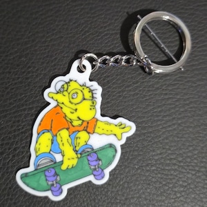 May include: A novelty keychain featuring a yellow cartoon character with glasses, wearing an orange shirt and blue shorts, skateboarding on a green board with purple wheels. The keychain has a silver metal ring and chain.