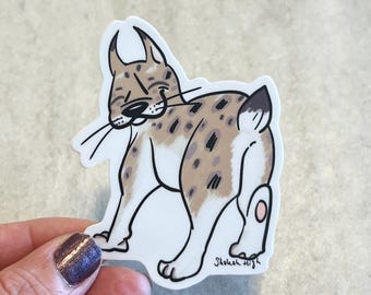 May include: A sticker featuring a cartoon lynx with a spotted coat in shades of brown and tan. The lynx is smiling and looking over its shoulder. The sticker has a white border and the artist's signature.