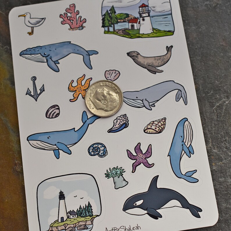 May include: A sheet of stickers featuring various ocean-themed illustrations, including whales, a sea lion, a lighthouse, coral, and seashells. The stickers are printed on white paper and are approximately 2.5 cm in diameter.