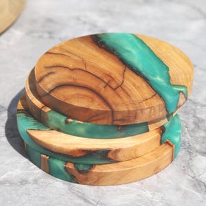 Może przedstawiać: Stos czterech okrągłych podkładek pod szklanki z jasnego drewna z turkusową żywicą. Drewno ma widoczne słoje. Turkusowa żywica tworzy uderzający kontrast. Każda podkładka ma średnicę około 10 cm.