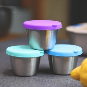 May include: Three small stainless steel containers with colorful lids. One container has a purple lid, another has a light blue lid, and the third has a blue lid. The containers are stacked on top of each other.