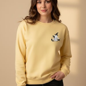 Peut inclure: Un sweat-shirt à col rond jaune pâle avec une broderie sur la poitrine gauche. Le motif représente un fantôme blanc portant un chapeau de sorcière noir, entouré de trois petits chats noirs dans différentes positions sur un fond clair.