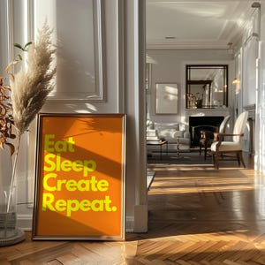 May include: A gold framed poster with an orange background and yellow text that reads "Eat Sleep Create Repeat."