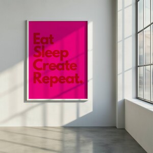 Peut inclure: Une affiche encadrée avec un fond rose vif et les mots "Eat Sleep Create Repeat" en rouge. L'œuvre est accrochée sur un mur blanc, avec des ombres naturelles.