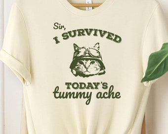 May include: A cream colored t-shirt with a green graphic of a cat wearing a helmet and goggles. The text on the shirt reads "Sir, I survived today's tummy ache."