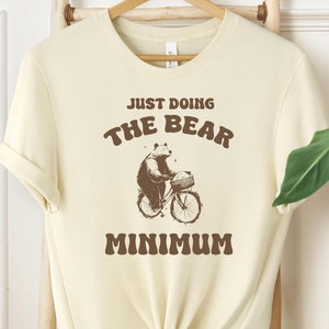 Puede incluir: Una camiseta de color crema con un oso marrón montando en bicicleta y el texto "Just Doing The Bear Minimum".
