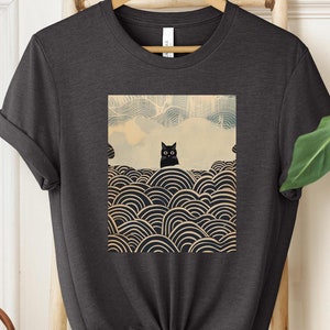 Puede incluir: Una camiseta gris oscuro con un gato negro sentado sobre un patr&oacute;n de c&iacute;rculos superpuestos. El gato mira directamente al espectador. El patr&oacute;n recuerda al arte japon&eacute;s.