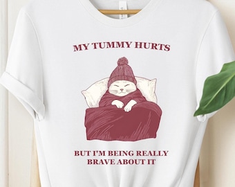 May include: White t-shirt with a graphic of a cat wearing a hat and a blanket, with the text "My tummy hurts but I'm being really brave about it."