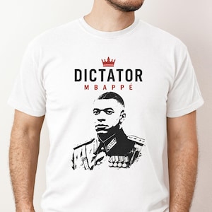 Può includere: Una t-shirt bianca a maniche corte con una grafica in bianco e nero di una persona in uniforme militare con medaglie. Sopra la grafica c'è un'icona a corona rossa e il testo "DICTATOR" in nero, con "MBAPPÉ" in lettere rosse sotto.
