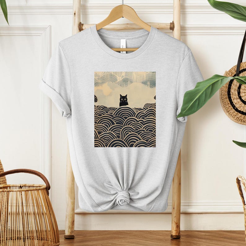 Puede incluir: Una camiseta blanca con un gato negro sentado sobre un fondo de olas blancas y negras. El gato tiene grandes ojos negros.