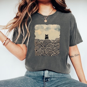 May include: Dark gray t-shirt featuring a graphic of a black cat in a landscape with stylized waves. The cat is centered in a rectangular frame with a light blue sky and a cityscape in the background. The shirt is a casual style.