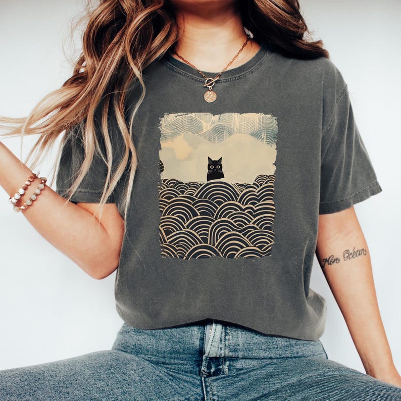 May include: Dark gray t-shirt featuring a graphic of a black cat in a landscape with stylized waves. The cat is centered in a rectangular frame with a light blue sky and a cityscape in the background. The shirt is a casual style.
