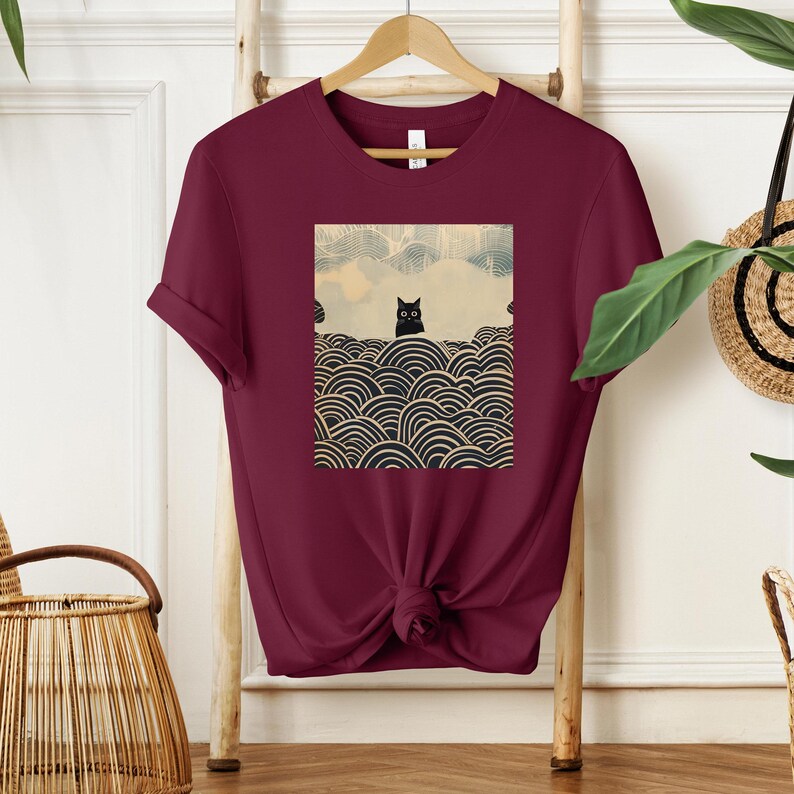 Puede incluir: Una camiseta color burdeos con un gato negro sentado sobre un fondo de olas estilizadas. El gato mira directamente al espectador.