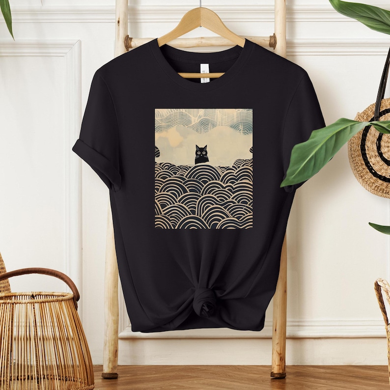 Puede incluir: Camiseta negra con un gr&aacute;fico de un gato negro sentado sobre un fondo estampado de olas beige y negras.