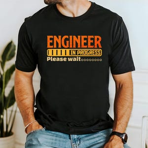 Może przedstawiać: Czarny T-shirt z tekstem "Engineer in progress" i grafiką paska postępu. Tekst "Please wait..." znajduje się pod grafiką.