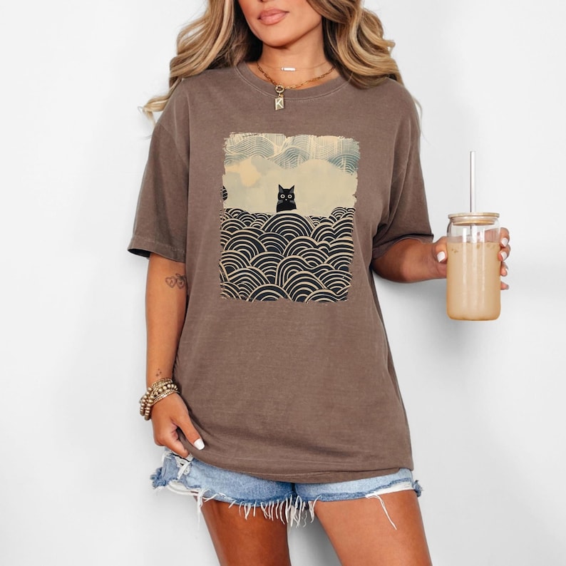 May include: Brown t-shirt featuring a graphic of a black cat in a landscape with stylized waves and clouds. The cat is centered in a rectangular design. The shirt is worn with denim shorts. A glass with a straw is held in the hand.