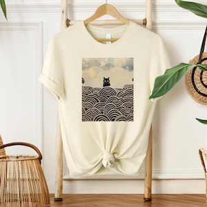 Puede incluir: Una camiseta de color crema con un gato negro sentado sobre un patr&oacute;n de olas en blanco y negro. El gato tiene grandes ojos negros.