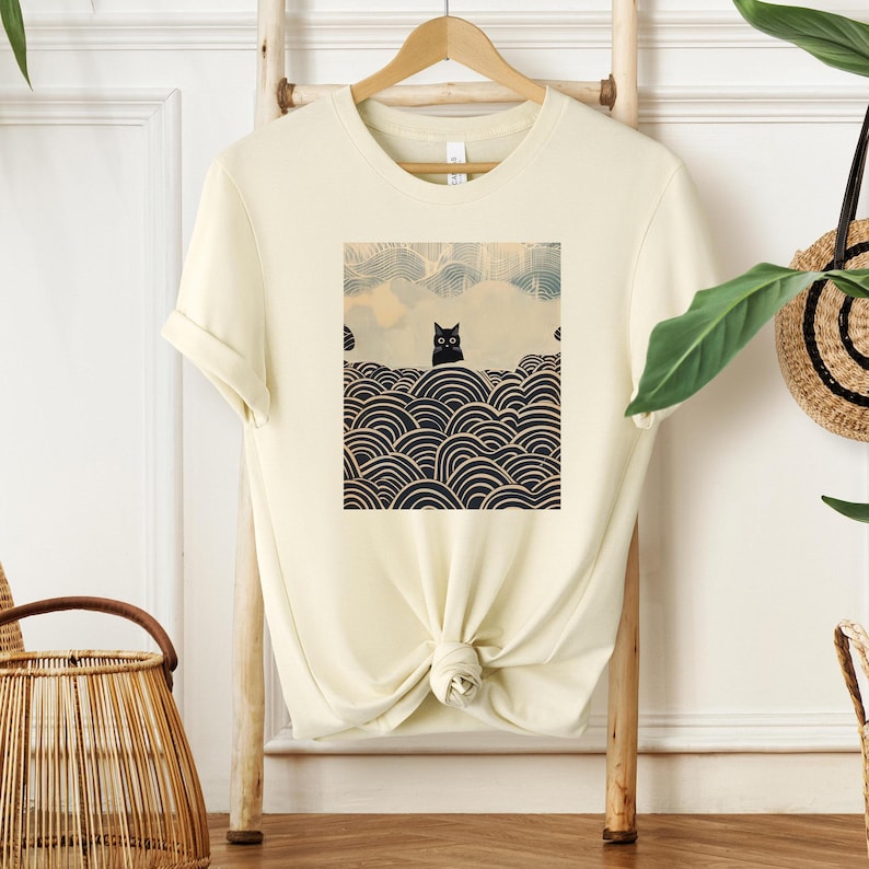 Puede incluir: Una camiseta de color crema con un gato negro sentado sobre un patr&oacute;n de olas en blanco y negro. El gato tiene grandes ojos negros.