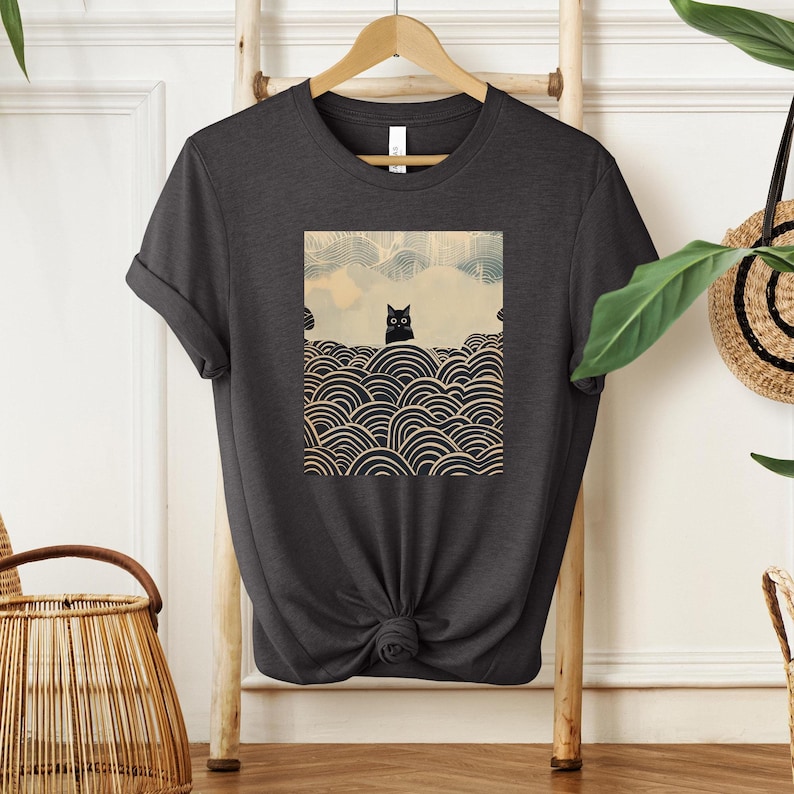 Puede incluir: Una camiseta gris oscuro con un gato negro sentado sobre un patr&oacute;n de c&iacute;rculos superpuestos. El gato mira directamente al espectador. El patr&oacute;n recuerda al arte japon&eacute;s.