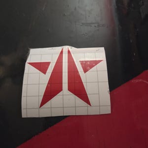 May include: A red vinyl decal sticker featuring a stylized star design split into two symmetrical halves. The decal is applied to a white backing paper printed with a light gray grid pattern to assist with alignment and application.