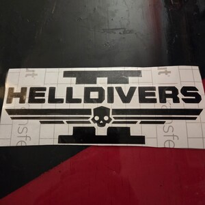 May include: A black vinyl decal featuring the text HELLDIVERS with a stylized skull icon centered below the word, flanked by horizontal wings, all set against a white grid-patterned transfer paper backing for application.