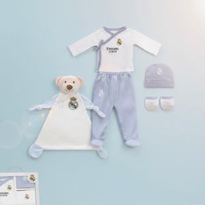 Puede incluir: Un conjunto de regalo para bebé con un conjunto blanco y azul claro con el logo del Real Madrid. El conjunto incluye una parte superior envolvente de manga larga, pantalones, un gorro y manoplas. También se incluye un oso de peluche y una mantita.
