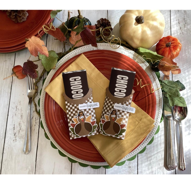 Puede incluir: Dos barras de chocolate envueltas en papel marr&oacute;n con un dise&ntilde;o de pavo y el texto "Choco" y "Feliz D&iacute;a de Acci&oacute;n de Gracias". Las barras de chocolate est&aacute;n sobre un mantel individual dorado y rojo con un tenedor y un cuchillo a un lado.
