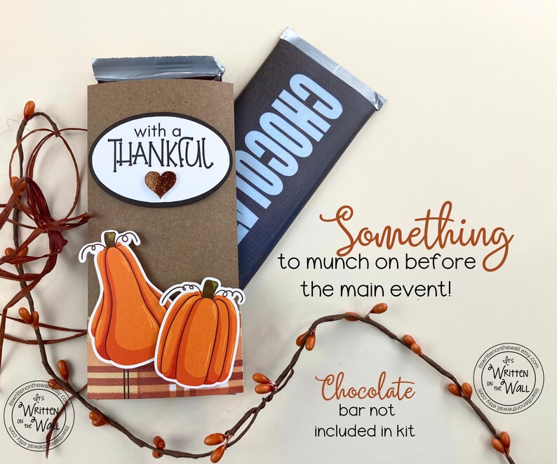 May include: A brown paper gift bag with a chocolate bar inside. The bag is decorated with a pumpkin and the text "with a thankful". The text "Something to munch on before the main event!" is written below the bag. The text "Chocolate bar not included in kit" is written below the text.