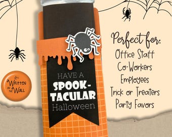 May include: A Halloween-themed gift holder with an orange and black color scheme. The holder features a black banner with the words "HAVE A SPOOK-TACULAR Halloween" in white. A decorative spider is attached to the holder.