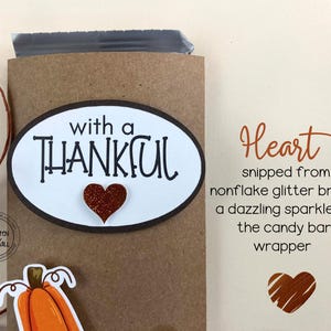 May include: A brown paper bag with a white oval sticker that says "with a THANKFUL" with a red glitter heart. The bag is decorated with a brown twine bow. The text "Heart snipped from nonflake glitter brings a dazzling sparkle to the candy bar wrapper" is written on a white background.