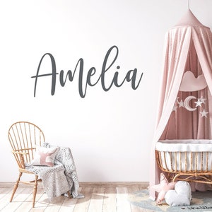 May include: A white wall with the name "Amelia" written in gray cursive script. A wicker crib with a pink canopy and a white cloud-shaped mobile hangs above it. A wicker chair with a white blanket and a pink pillow sits in the foreground.