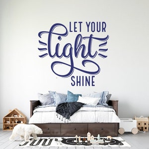Puede incluir: Una calcomanía de pared azul marino con el texto "Let your light shine" en una fuente de escritura decorativa. La calcomanía se muestra sobre un marco de cama de madera con ropa de cama blanca y una manta negra.
