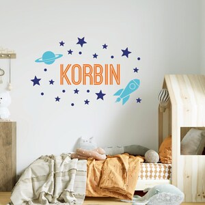 Può includere: Un adesivo da parete blu e arancione con il nome "KORBIN" in lettere arancioni, un razzo, un pianeta e stelle. L'adesivo è su una parete bianca sopra un letto con una coperta bianca e cuscini arancioni.