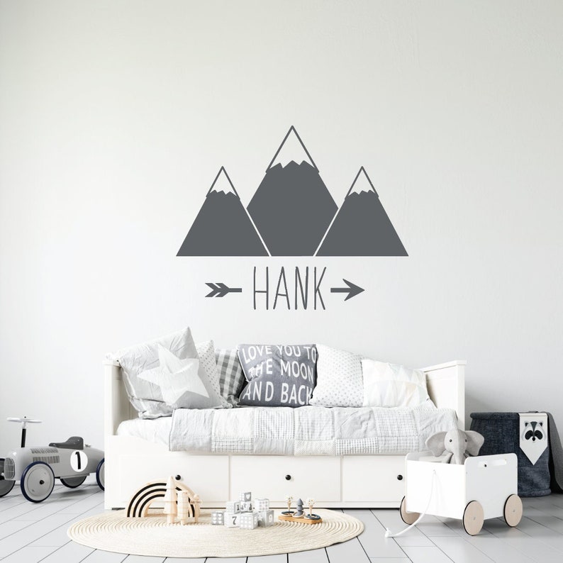 May include: A gray mountain range wall decal with the name "HANK" in gray text with arrows on either side. The decal is on a white wall above a white daybed with gray and white patterned pillows.