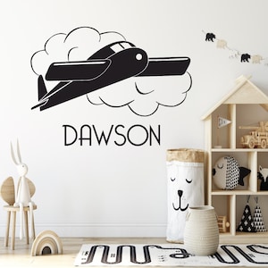 Puede incluir: Adhesivo de pared de avión negro con el nombre "DAWSON". El avión vuela a través de una nube. Ideal para la habitación de un niño o guardería.