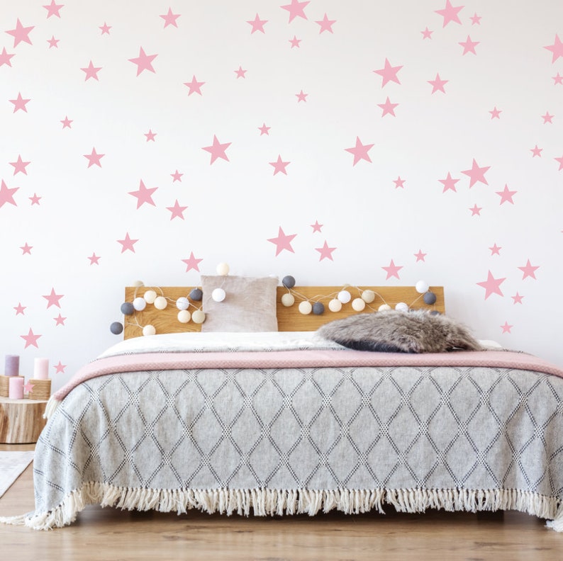 May include: A white wall with pink star stickers, a bed with a gray blanket and a pink throw, and a wooden headboard with a string of lights.